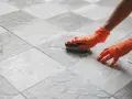 Mani con guanti arancioni puliscono le fughe del pavimento con una spazzola, mostrando come pulire le fughe del pavimento.