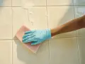 Main tenant, une main gantée de bleu frotte le carrelage de la salle de bain avec une éponge rose, faisant mousser le savon.