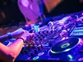 Hände am DJ-Pult, die die Regler für verschiedene techno genres bedienen. Neonlicht und pulsierende Beats.