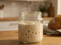 Blubbernder Sauerteig im Glas, der nach Essig riecht, dampft leicht. Daneben liegt ein frisch gebackenes Brot.