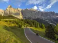 Un motociclista percorre una strada panoramica tra prati verdi e boschi di conifere, con le maestose Dolomiti sullo sfondo. Un'avventura indimenticabile in moto.