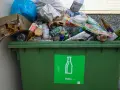 Przepełniony zielony kontener na śmieci z butelkami i szkłem, z zieloną naklejką "Szkło".