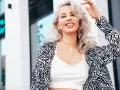 Uśmiechnięta blondynka z falowanymi włosami, efekt farbowania włosów na blond. Wzorzysta marynarka i biały top.
