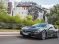 Czarny BMW i8 pędzi ulicą, mijając nowoczesne budynki i zielone drzewa.