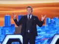 Rafał Brzozowski w garniturze, z rękami uniesionymi na boki, prowadzi program "Jaka to melodia?".
