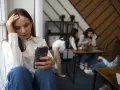 Dziewczyna z telefonem w dłoni, zmartwiona, siedzi przy oknie, podczas gdy inni są w tle.