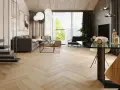 Nowoczesny salon z szarą sofą, szklanym stolikiem i jadalnią. Wzrok przyciągają panele bez dylatacji w progu, tworzące jednolitą, drewnianą podłogę w jodełkę.