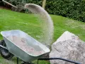 Schubkarre mit Sand, während eine Schaufel Sand auf den Rasen rieselt.