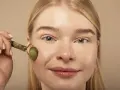 Kobieta z blond włosami używa jadeitowego masażera do twarzy, wspierając poprawę owalu twarzy domowymi sposobami.