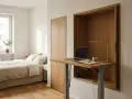 Minimalistyczne biurko inspiracje w sypialni. Drewniana wnęka skrywa regulowany blat z laptopem, lampką i notatnikiem. Obok łóżko i szafa.