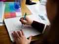 Ilustrator pracuje nad projektem, zamieniając litery na krzywe. Na stole leżą przybory do rysowania i próbnik kolorów.
