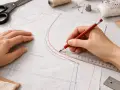 Main tenant une règle et un crayon pour comment agrandir un patron de couture. Des ciseaux, du fil et des épingles sont visibles.