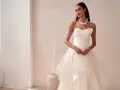 Elegancka panna młoda w sukni ślubnej z falbanami, prezentująca najmodniejsze suknie ślubne. Delikatny naszyjnik z pereł podkreśla jej urodę.