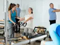 Frauen und Männer trainieren im **Reha-Sport** auf Pilatesgeräten. Eine Trainerin hilft einer älteren Frau, während ein Mann mit einem Ball übt.