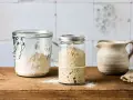 Zwei Gläser mit Sauerteig Weizen, eines mit aktivem, blasigem Sauerteig, das andere mit trockenem Ansatz. Daneben ein kleiner Krug.