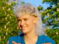 Persona sonriente con un moderno mullet rubio, aretes y un suéter azul con rayas naranjas, frente a un árbol en flor.