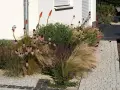 Blumenbeet an Hauswand: Feuchtigkeit durch Wasserhahn und Bewässerungssystem. Ziergräser, Sonnenhut und Fackellilien blühen üppig.
