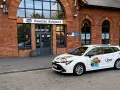 Biały samochód Ubera zaparkowany przed dworcem kolejowym. Kierowca z własnym samochodem czeka na pasażerów.