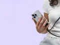 Dłoń trzyma telefon z przezroczystym etui i magnetycznym mocowaniem. Czarny brelok do telefonu przewieszony przez ramię.