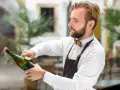 Barman w białej koszuli i brązowym fartuchu pokazuje, jak otwierać szampana, trzymając butelkę i wyciągając korek.
