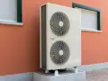 Unità esterna di una pompa di calore, con due ventole, montata su una base rialzata contro un muro arancione con finestre.