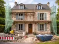Façade maison en pierre en rénovation, échafaudages et bâches vertes protègent les travaux. Matériaux de construction et benne à gravats au premier plan.
