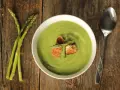 Ciotola di vellutata verde brillante, guarnita con crostini e asparagi freschi. Accanto, un mazzetto di asparagi e un cucchiaio. Un delizioso pesto di asparagi pronto da gustare.