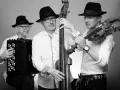 Trzech muzyków w czarnych kapeluszach grających muzykę klezmerską: akordeonista, basista i skrzypek.