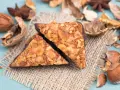 Zwei vegane Nussecken mit Schokoladenrand, bedeckt mit gehackten Nüssen, liegen auf einem Jutesack, umgeben von verschiedenen Nüssen und Sternanis.