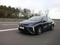 Ciemnoniebieski samochód na wodór Toyota Mirai jedzie po drodze. Na drzwiach widoczny napis "Napęd Prąd Wodór".