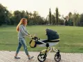 Ruda kobieta z uśmiechem pcha wózek podczas pierwszego spaceru z noworodkiem w parku.