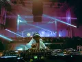 Un DJ année 2000, lunettes de soleil et bandeau, mixe sous des lasers bleus et violets devant une foule en délire.