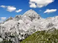 Majestätischer Triglav im Sonnenschein, bereit für die Triglav-Besteigung. Grüne Hänge und Felsen unter blauem Himmel mit Wolken.