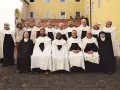 Gruppe von unbeschuhten Karmeliten in braunen Kutten und weißen Umhängen posiert vor einem gelben Gebäude.