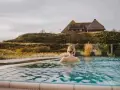Frau genießt entspannten Wellness Tag in Schleswig-Holstein. Ein idyllischer Pool mit Blick auf Reetdachhaus und Dünenlandschaft.