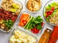 Gotowe pojemniki z jedzeniem na piknik: ryż z fasolą i pieczarkami, warzywa pokrojone w słupki, kalafior, sałatka z kiełkami i pomidorami, hummus i napoje.