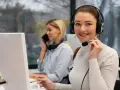 Uśmiechnięta key account manager w słuchawkach z mikrofonem pracuje przy komputerze. W tle koleżanka rozmawia przez telefon.