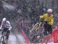 Cyclists battle snowy conditions. Wout van Aert, in yellow, appears to be recovering from a crash, while another rider in white pushes on.