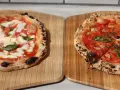 Zwei frisch gebackene Pizzen auf Holzbrettern, eine Margherita, die andere mit Tomaten und Basilikum.