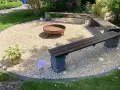 Gemütliche Sitzecke im Garten mit Kiesboden, Feuerstelle und Holzbänken. Perfekt für laue Sommerabende.