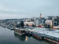 Widok na zimowe miasto nad rzeką z łodziami i ośnieżonymi budynkami.