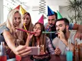Grupa przyjaciół świętuje urodziny mężczyzny, dmuchając w trąbki i robiąc selfie przy torcie.