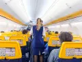 Wnętrze samolotu Ryanair, stewardessa obsługuje pasażerów. Żółte zagłówki siedzeń i pomarańczowe pasy na suficie tworzą słoneczną atmosferę.