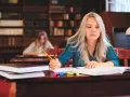 Kobieta z długimi blond włosami pisze w zeszycie, siedząc przy biurku w bibliotece.