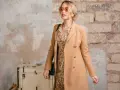 Elegancka kobieta w płaszczu w camelowy kolor, kwiecistej sukience i okularach przeciwsłonecznych, obok starej skrzyni.
