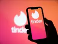 Dłoń trzyma smartfon z logo Tinder. W tle widać większe, rozmyte logo aplikacji, sugerujące nowe tinder powiadomienia.