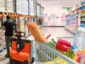 W magazynie FMCG, pracownik obsługuje wózek widłowy, podczas gdy wózek sklepowy pełen produktów czeka na zakupy.