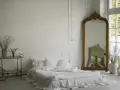 Elegancka sypialnia z łóżkiem w bieli, ozdobiona sztukaterią przypominającą lamperię angielską. Złote lustro i wózek dodają blasku.