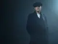 Mężczyzna w kaszkiecie i płaszczu, z charakterystycznym wąsem, emanuje chłodnym spojrzeniem w stylu Peaky Blinders.