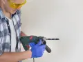 Man in safety glasses and blue gloves holding a drill to a white surface.